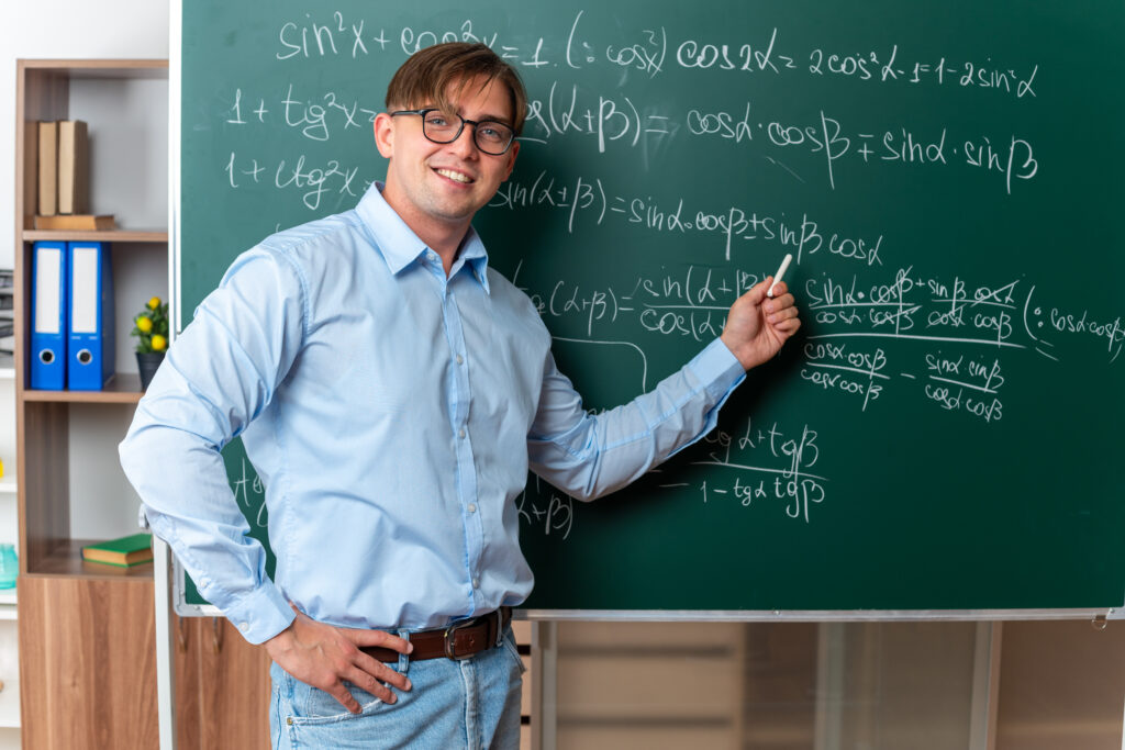 young male teacher wearing glasses holding chalk explaining lesson standing near blackboard with mathematical formulas in classroom